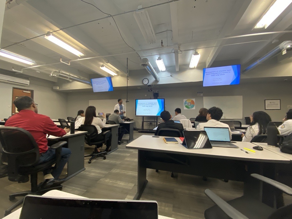 A classroom setting at Larkin University College of Pharmacy where a PharmD student presents research on inhalable lipid-based drug delivery for lung cancer. Faculty and students listen attentively, with laptops and notebooks open.