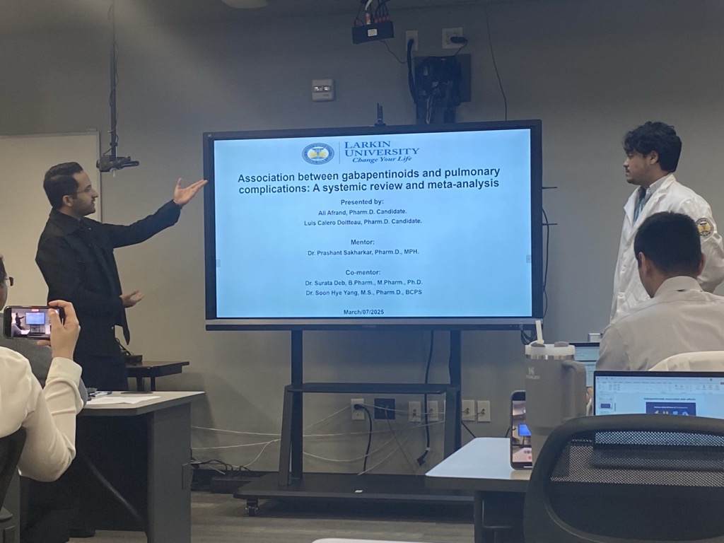 Two PharmD students at Larkin University presenting a research project on gabapentinoids and pulmonary complications in a classroom, with a projected slide displaying their study title and mentor names.