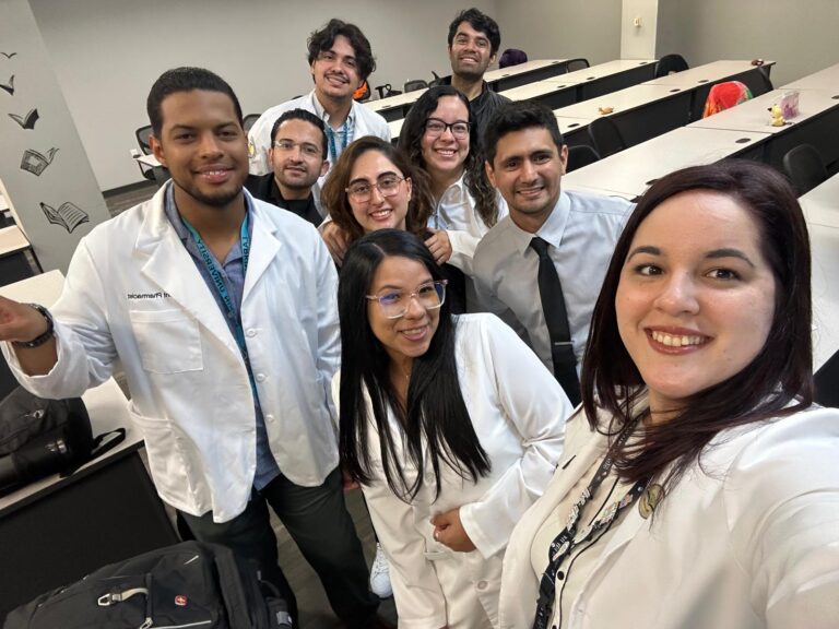 Smiling PY1 PharmD students from Larkin University College of Pharmacy take a selfie in a classroom while wearing lab coats after presenting their research projects.