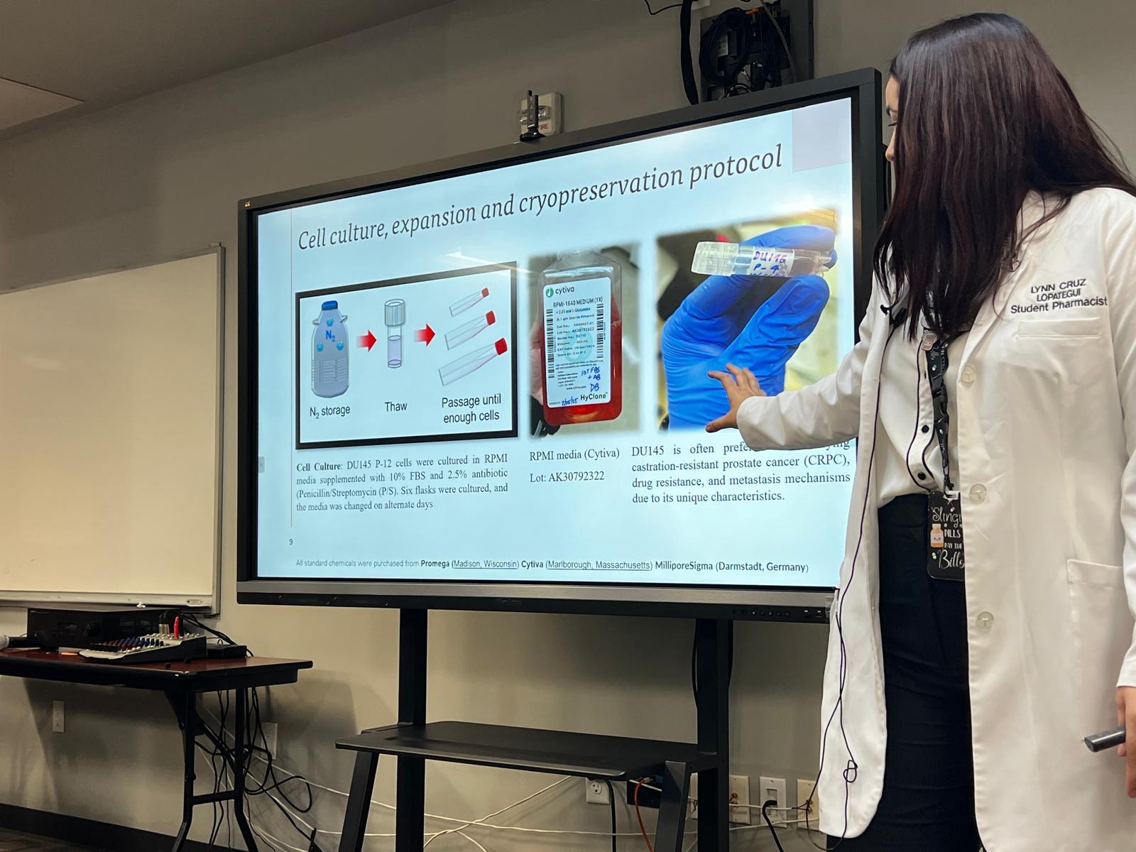 PharmD student in a lab coat presenting research on cell culture, expansion, and cryopreservation protocols in front of a classroom screen.